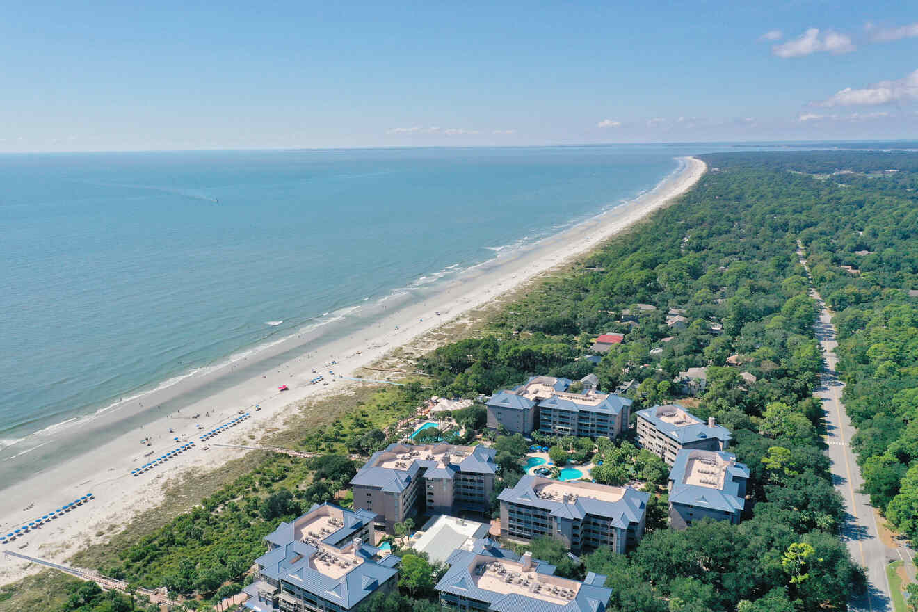 Aerial view of a coastal resort with beachfront property, multiple buildings, pools, and a long stretch of beach bordered by a forest on one side and the ocean on the other.