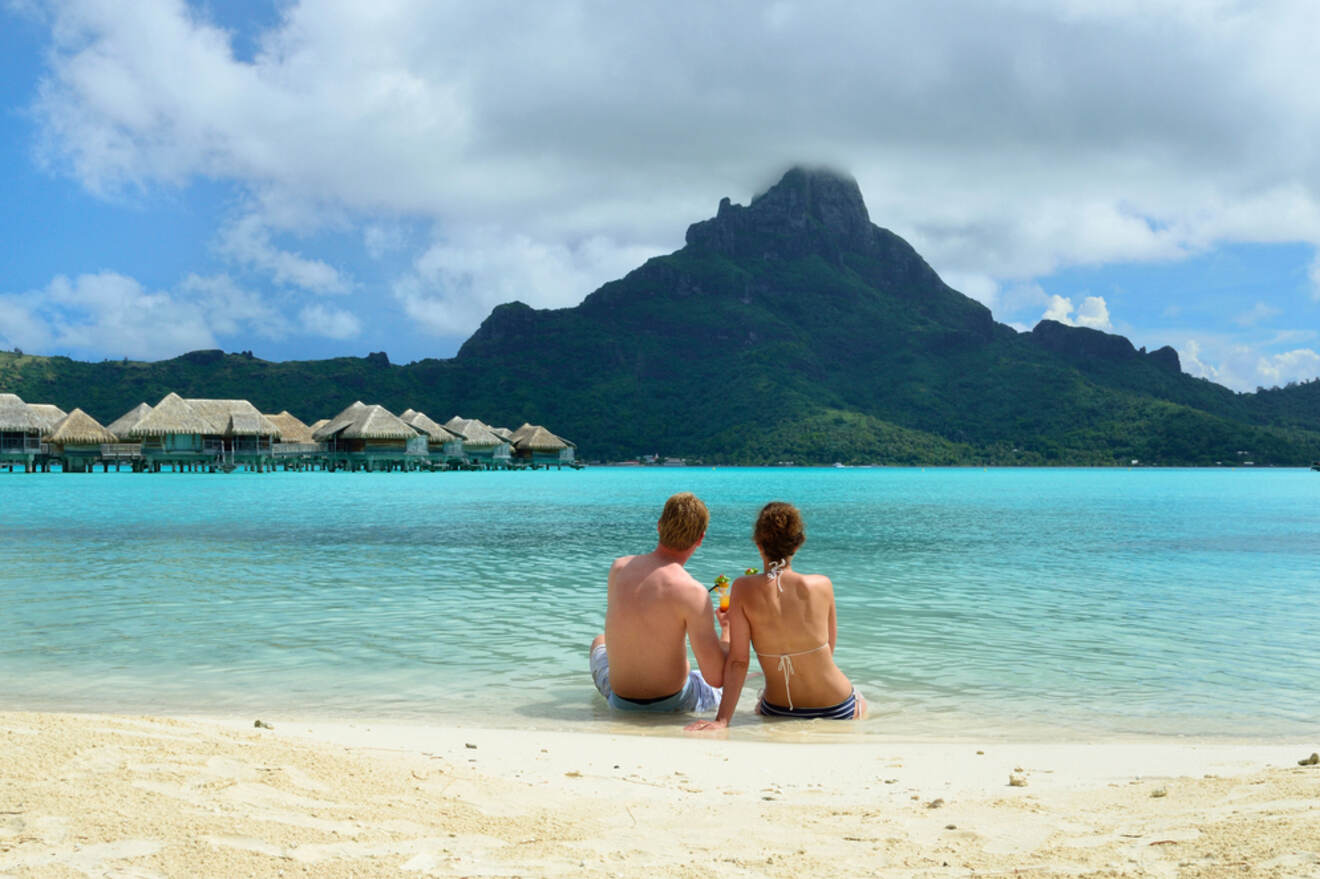 Two people sit on a sandy beach facing clear blue water, with overwater bungalows and a mountainous island in the background.