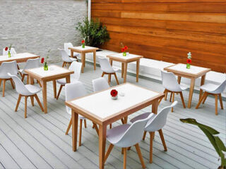 Outdoor seating area with wooden tables and white chairs, featuring single red flowers in small vases on each table. Wooden panel wall and stone wall in the background with some greenery.