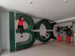 Two people are posing on a large 3D "BCG" sculpture indoors. One person is sitting on the letter "B," and the other is lying down inside the letter "O.