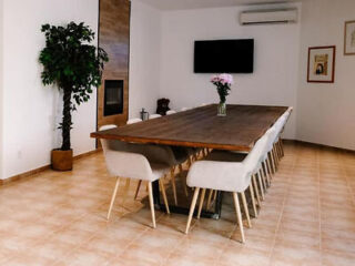 A spacious dining room with a long wooden table, light-colored chairs, and a TV mounted on the wall