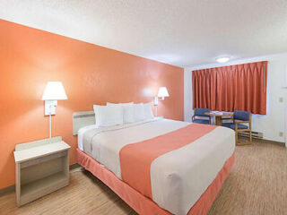 A hotel room with a king-size bed, two side tables with lamps, an orange accent wall, a window with an orange curtain, and a small table with two chairs.