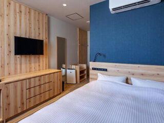 Hotel room featuring a double bed with a wooden headboard, a wall-mounted TV, wooden furniture, a mini fridge, and an air conditioning unit. The decor is modern with blue and wood accents.