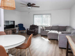 A living room with a gray sectional sofa, a brown recliner, a blue chair, a TV on a stand, a round dining table with four chairs, wood flooring, a ceiling fan, and a window with blinds.