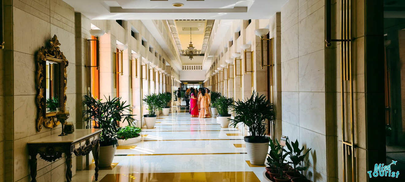 A luxurious hallway lined with plants and ornate mirrors inside a hotel, with people in colorful traditional attire in the distance.