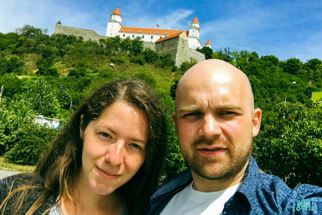 author of the post with her husband stand in front of grassy trees with a large hill and an old castle with red roofs on top, under a blue sky.