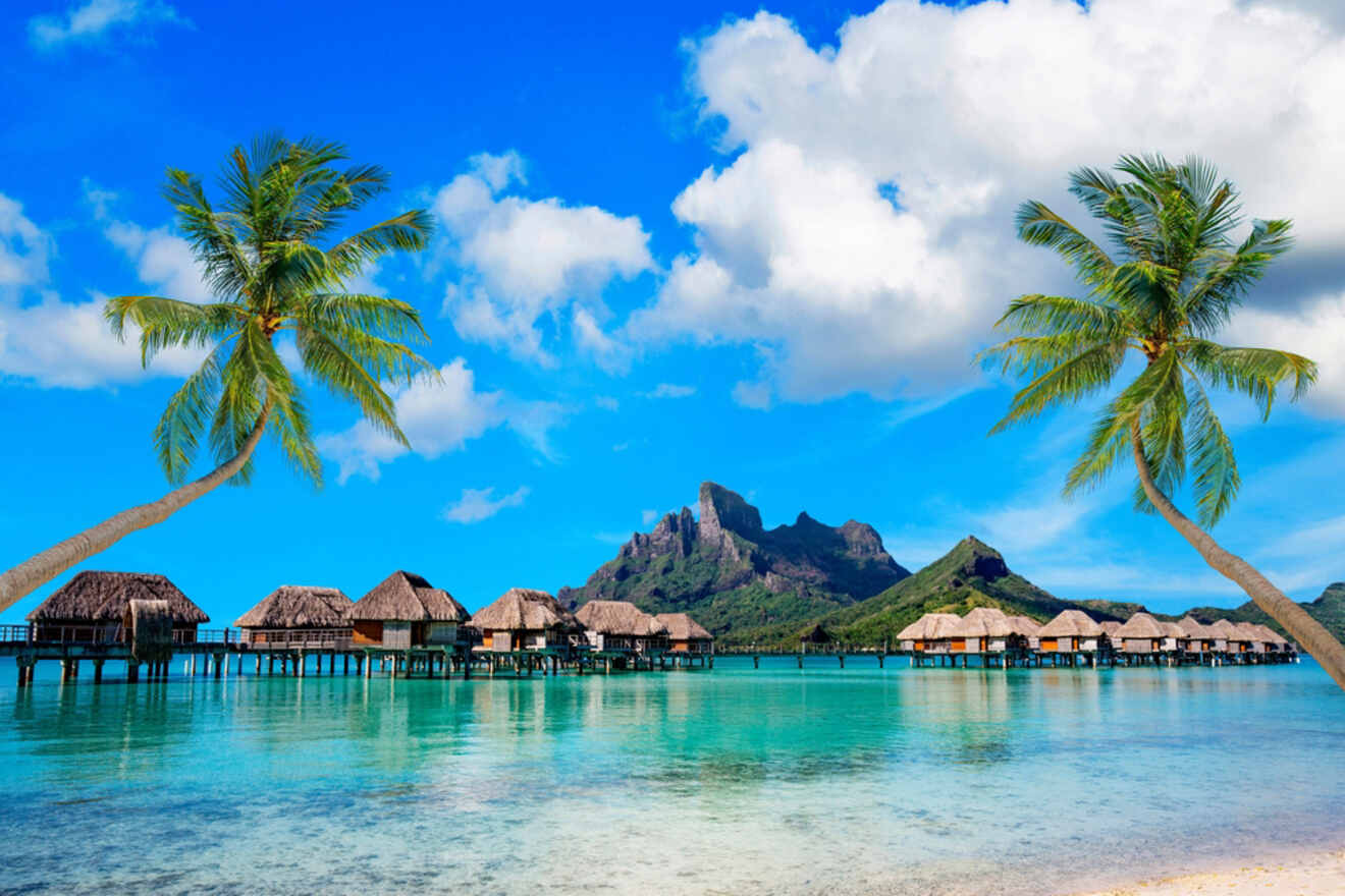 A picturesque tropical scene with two palm trees framing overwater bungalows, set against a backdrop of a clear blue sky and the dramatic silhouette of a mountain rising from the island.