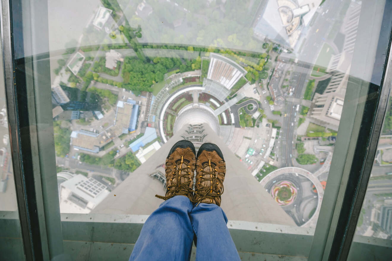 Looking down from a glass floor observation deck, a person wearing hiking boots and jeans peers over a cityscape below. The view captures buildings, roads, and green spaces from a significant height.