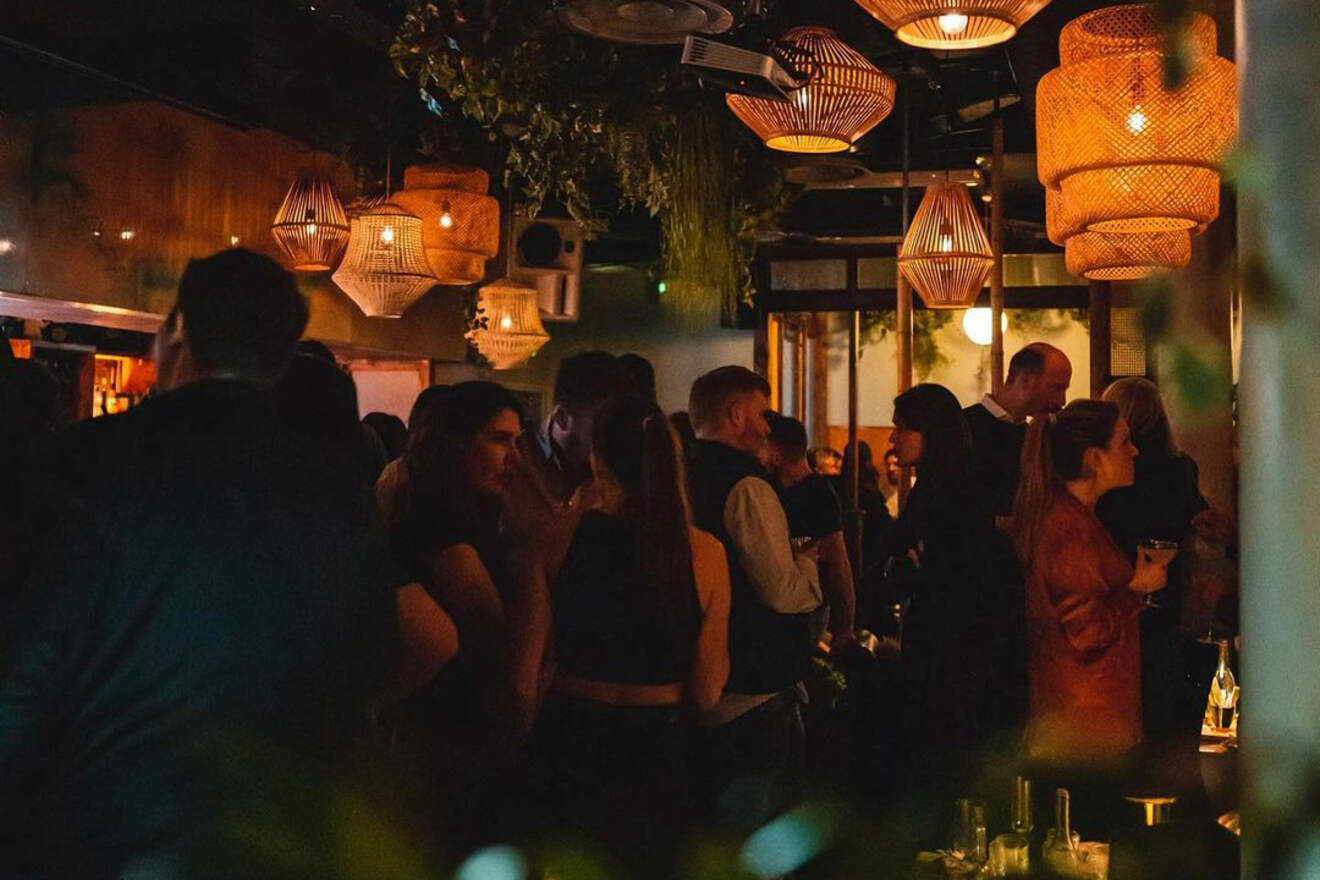 A dimly lit cocktail bar with hanging woven lamps, lush greenery, and a crowd of people engaged in conversation.