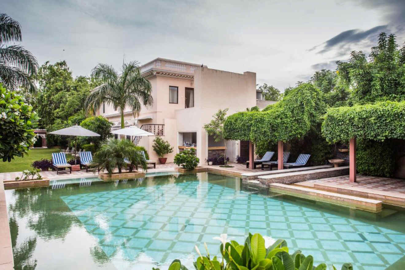 A large, luxurious villa with a swimming pool and loungers surrounded by greenery and trees under a cloudy sky.