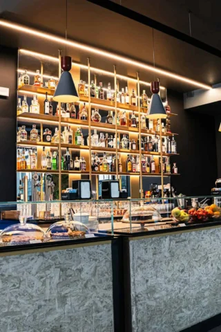 A modern bar with a well-stocked liquor shelf, hanging lights, glass showcases with pastries, and a fruit display on the counter.