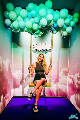 Kathryn, team member of Misstourist, sitting on a swing adorned with pastel balloons in a decorative room with a theme of clouds and sky.