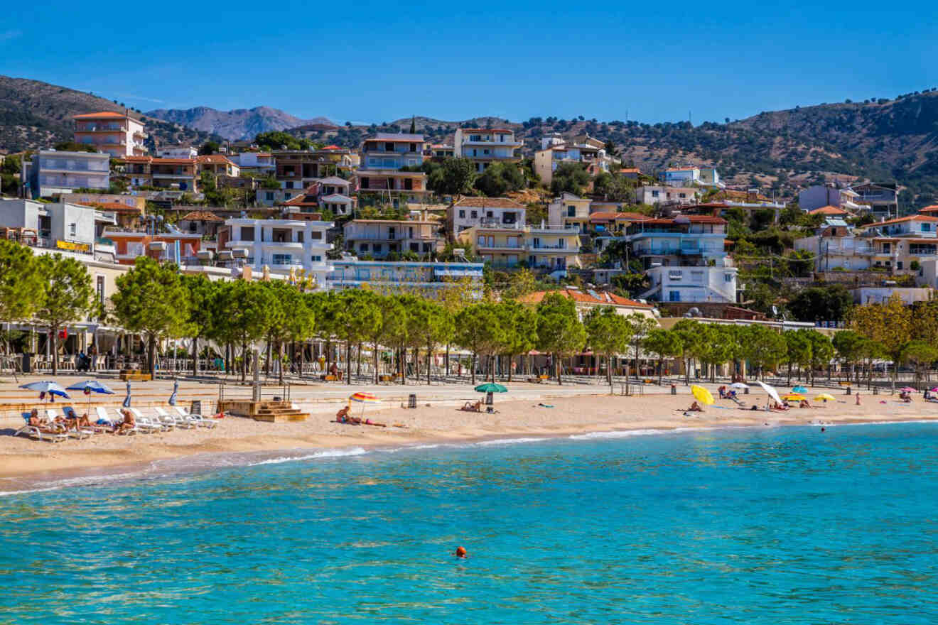 Himarë, Albania, viewed from the sea, with a beach lined with umbrellas and people relaxing, and the town's buildings rising up on the hillside against a backdrop of mountains.