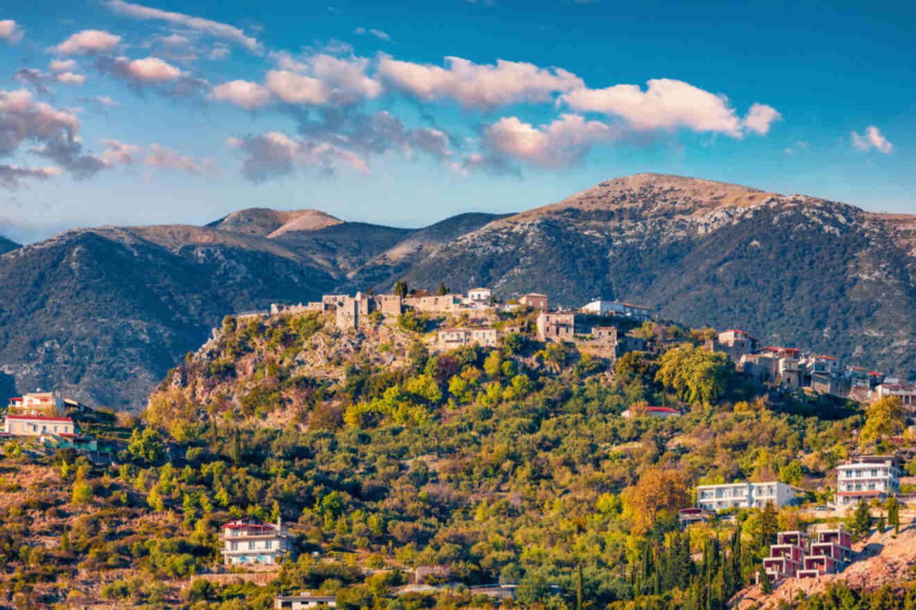 A view of the ruins of a fortress atop a hill in Albania, surrounded by lush greenery and residential homes, with mountains in the background under a bright blue sky.