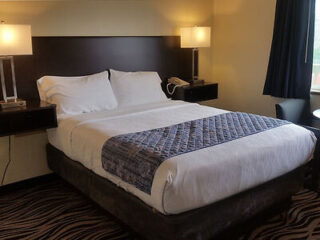 A neatly made bed with white linens and a patterned throw blanket in a hotel room, flanked by two lamps on bedside tables, a telephone, and a carpeted floor with a window to the right.