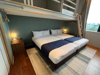 A modern bedroom with two single beds pushed together, nightstands with lamps, a ladder leading to bunk beds above, and a window with a city view. Rug on wooden floor.