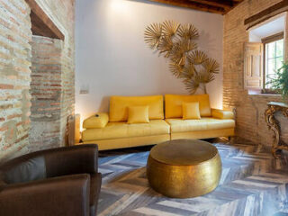 A living room features a yellow sofa with four cushions, a brown armchair, a round golden coffee table, herringbone patterned flooring, a brick wall, and a window with wooden shutters.