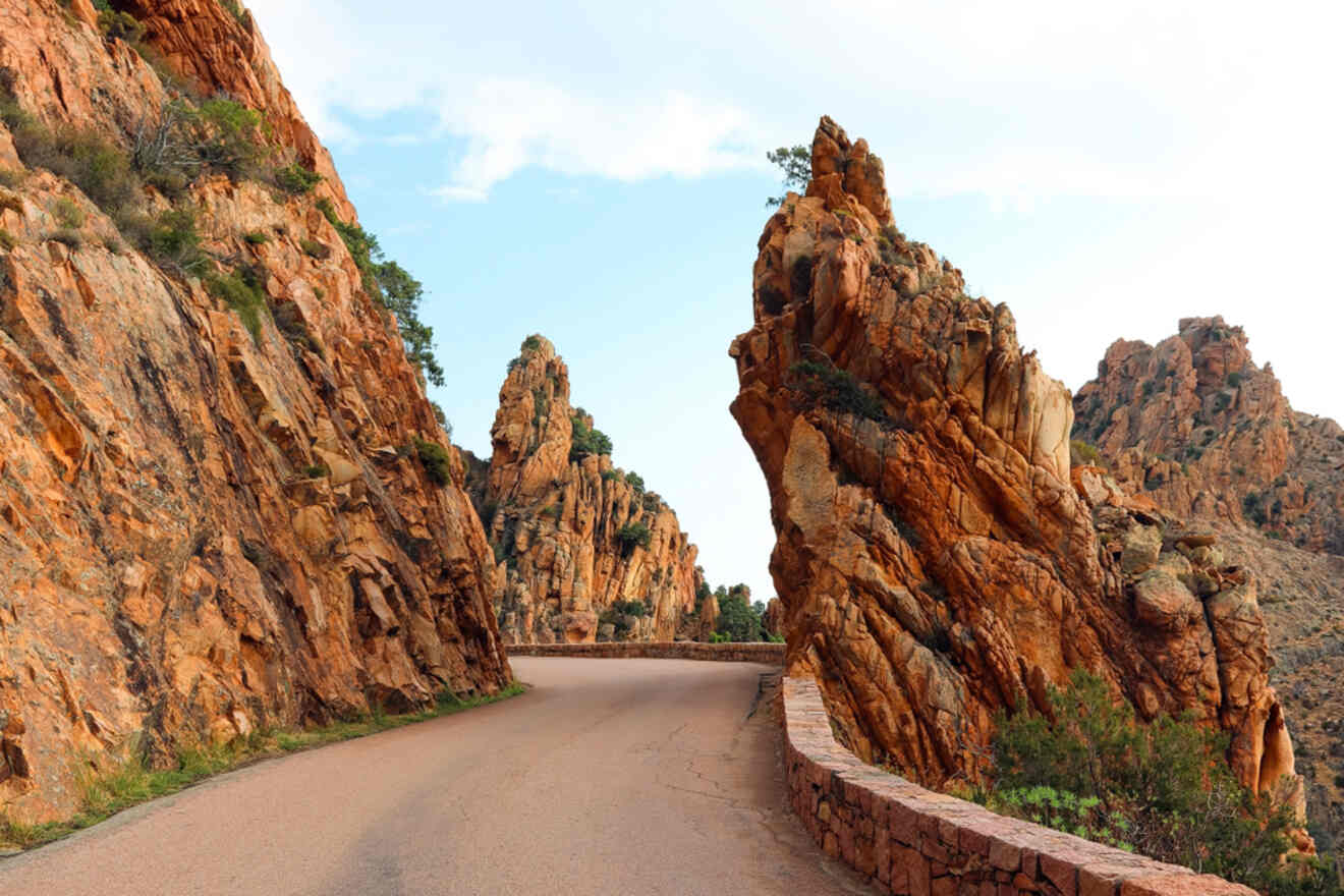 A scenic mountain road winding through jagged, towering red rock formations, with a low stone wall lining the path.