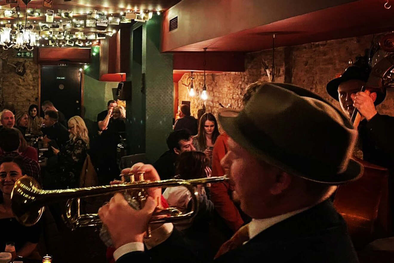 A jazz musician in a hat plays trumpet as a band performs for a lively audience in a cozy, warmly lit bar.