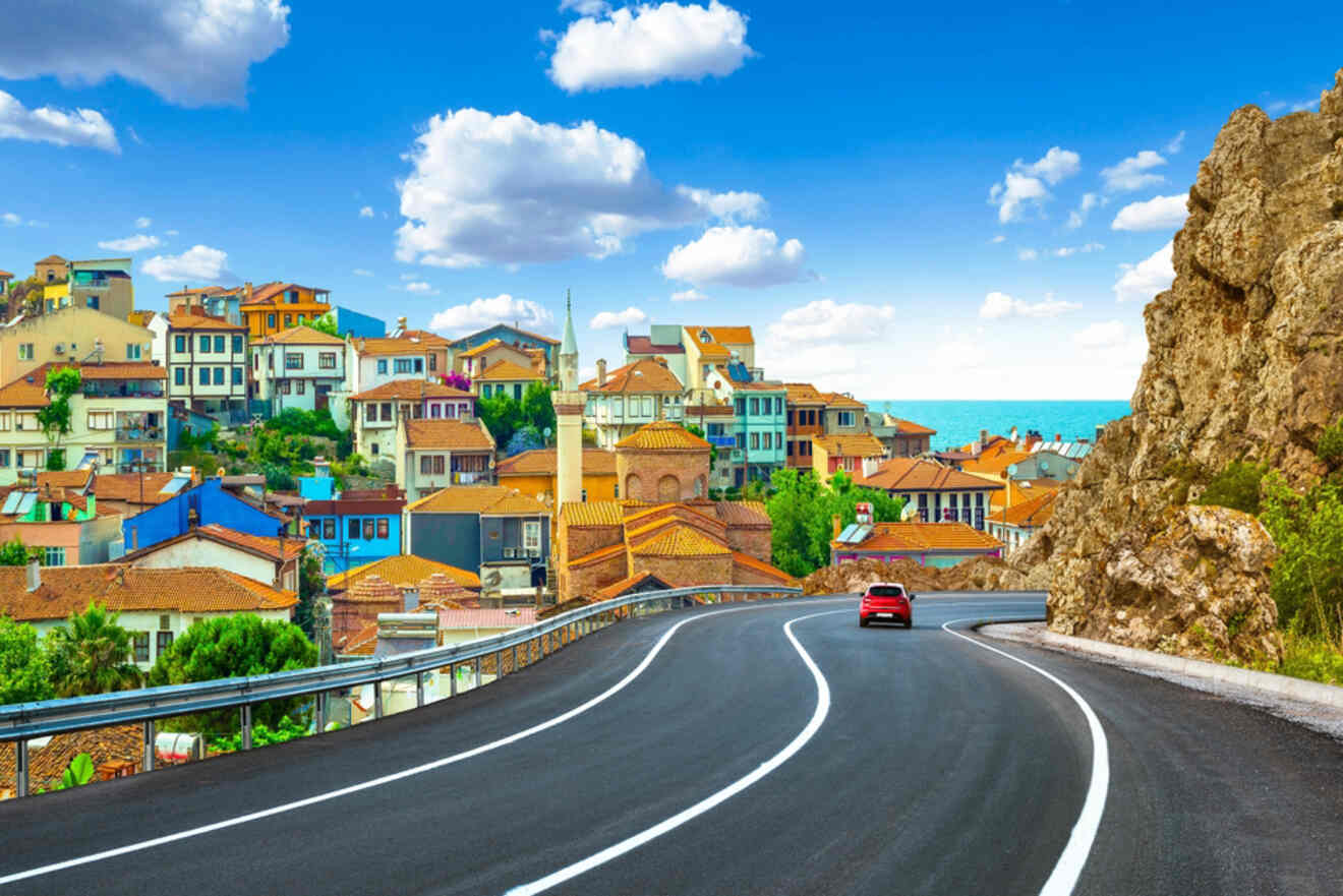 A winding road leads to a coastal town with colorful buildings under a bright blue sky with scattered clouds. A red car is driving along the road. Rocky cliffs are visible to the right.