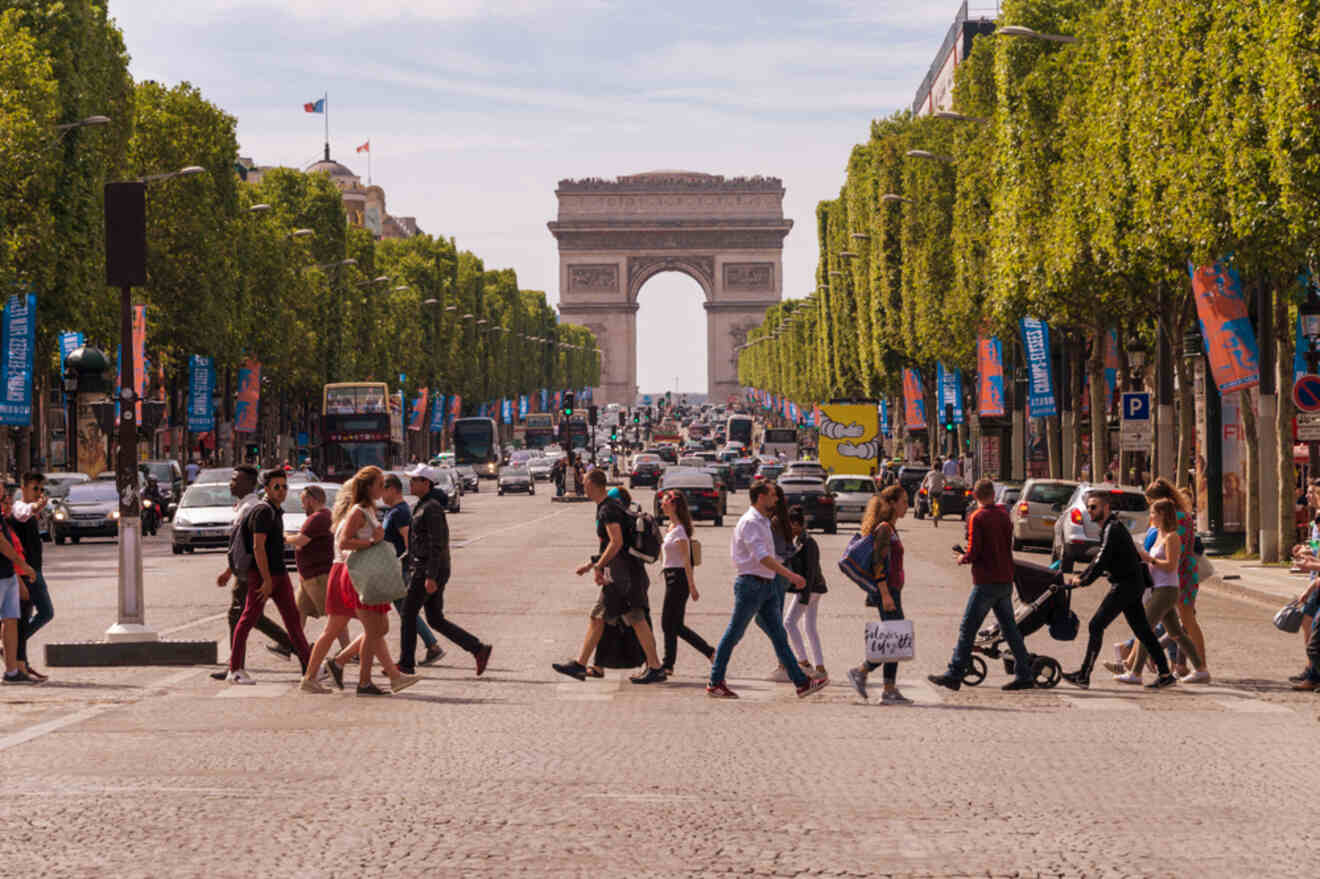 People cross a busy street in Paris with the Arc de Triomphe in the background, surrounded by trees and buildings.