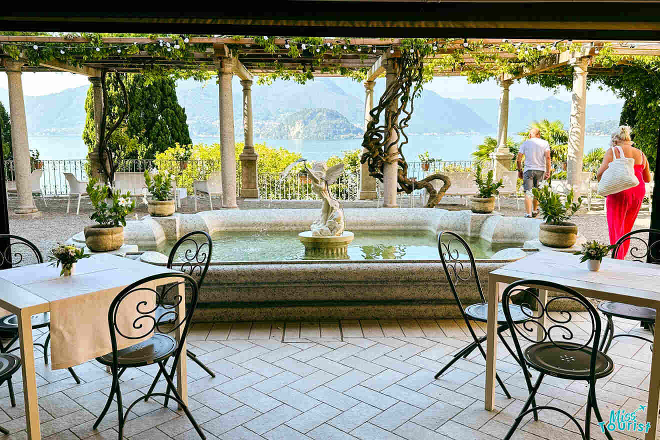 Outdoor dining area with iron chairs and tables. Overlooking a small fountain with a statue centerpiece. Two people in the background near lake and mountains. Greenery surrounds the area.