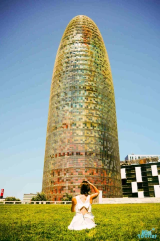 author of the post sits on grass facing a tall, cylindrical building with a colorful, mosaic-like exterior under a clear blue sky.