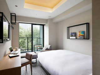 Modern hotel room with a large bed, a desk with a laptop, a chair, a wall-mounted TV, and a window with a balcony overlooking trees. Two framed pictures hang on the wall above the bed.