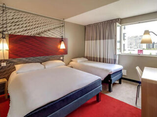 A hotel room with two beds, a desk, chair, modern light fixtures, patterned headboard, and a window with curtains partially open, offering a view of buildings outside.