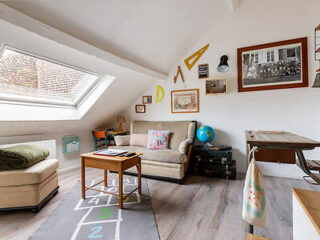 Attic room with a skylight, cozy sitting area, hopscotch rug, and various framed pictures and decorations on the walls. There is a small desk, a globe, and a mix of modern and vintage furniture.