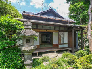 A traditional Japanese house with sliding doors, wooden exterior, a stone lantern in the garden, and greenery surrounding the area.