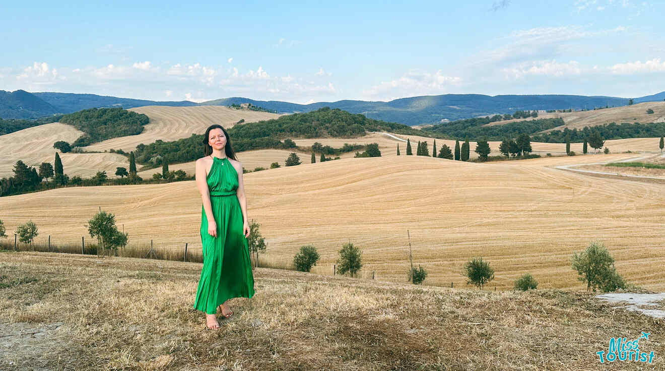 The writer of the post stands in a flowing green dress on a ridge overlooking vast Tuscan hills with scattered trees and farm fields.