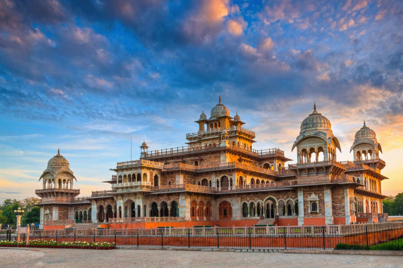 A large, ornate building with multiple domes and intricate architectural details stands under a dramatic sky with scattered clouds. A fence surrounds the structure.