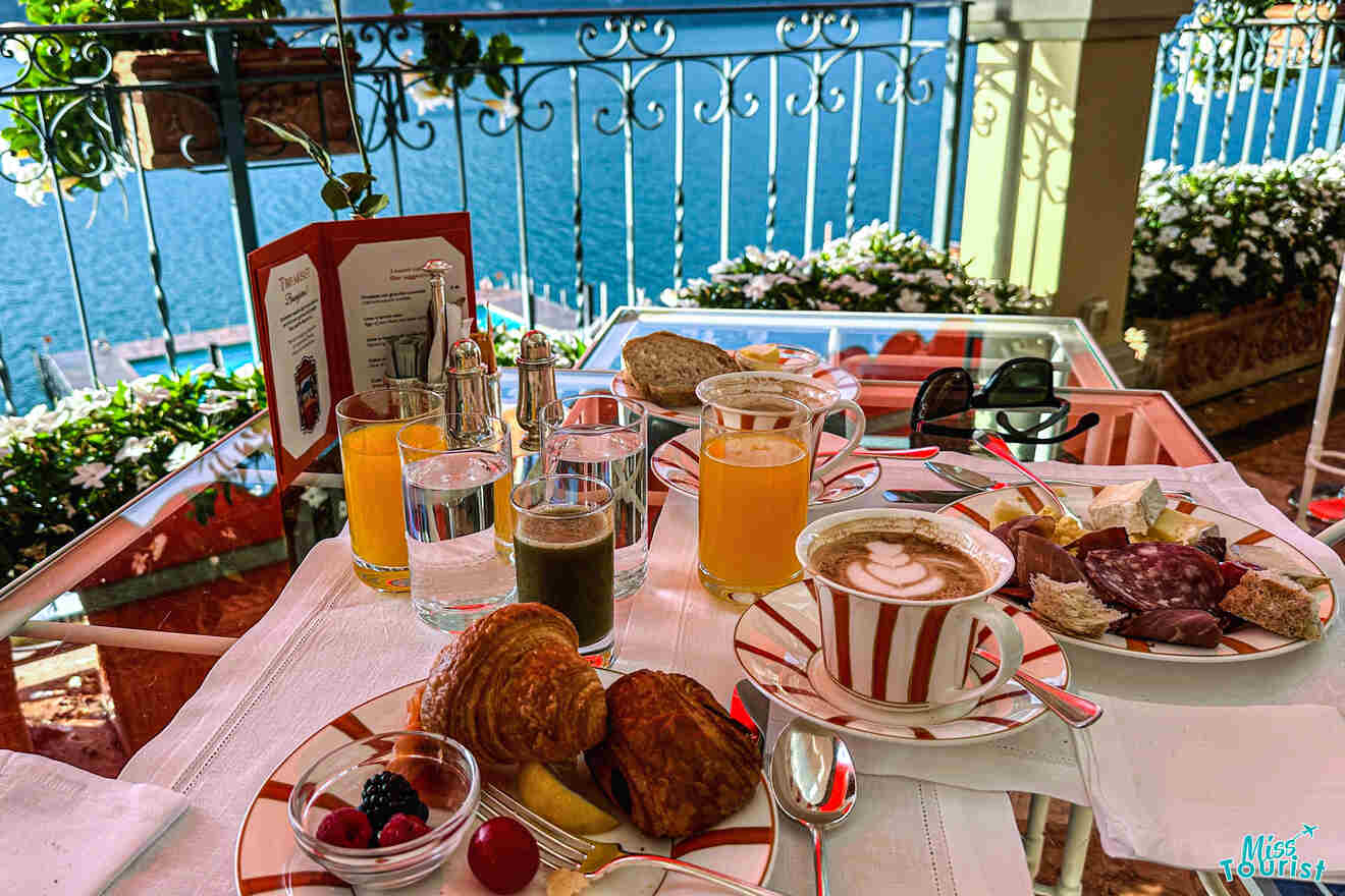 A breakfast table on a balcony overlooking water, featuring croissants, fruit, juice, coffee, and cold cuts. The table is set with red-and-white plates and glasses.