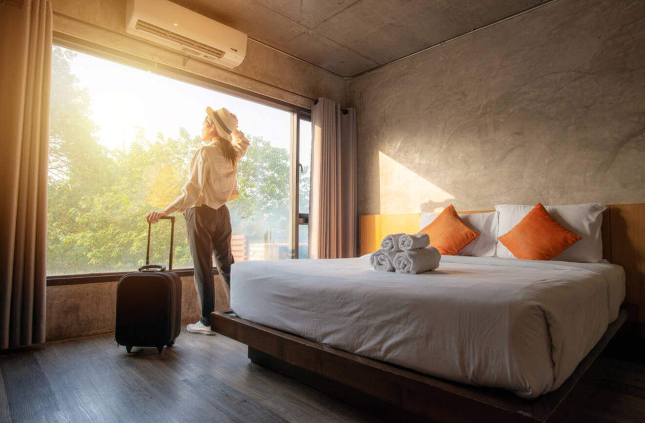 A person with a suitcase looks out of a large window in a hotel room with a neatly made bed and three orange cushions. Sunlight streams in, illuminating the room.