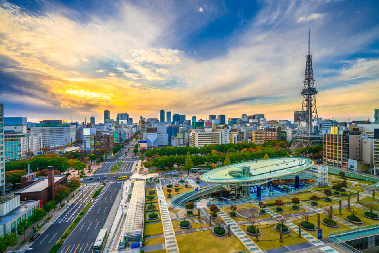 Aerial view of an urban park with modern buildings, trees, and a tall tower in the background at sunset. The area features a curved structure and city streets with light traffic.