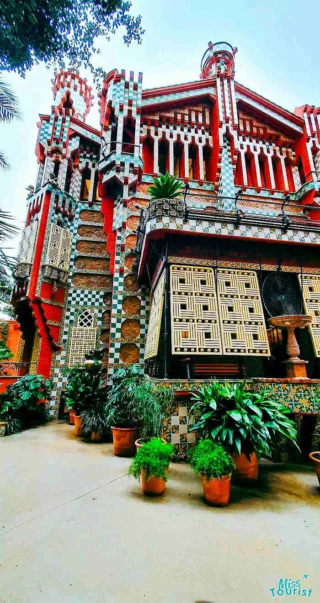 A brightly colored, intricately designed building with various geometric patterns and lush greenery in pots around its base.