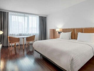 Modern hotel room with a large bed, two chairs and a table near a window with sheer curtains, wooden floor, and minimal decor.