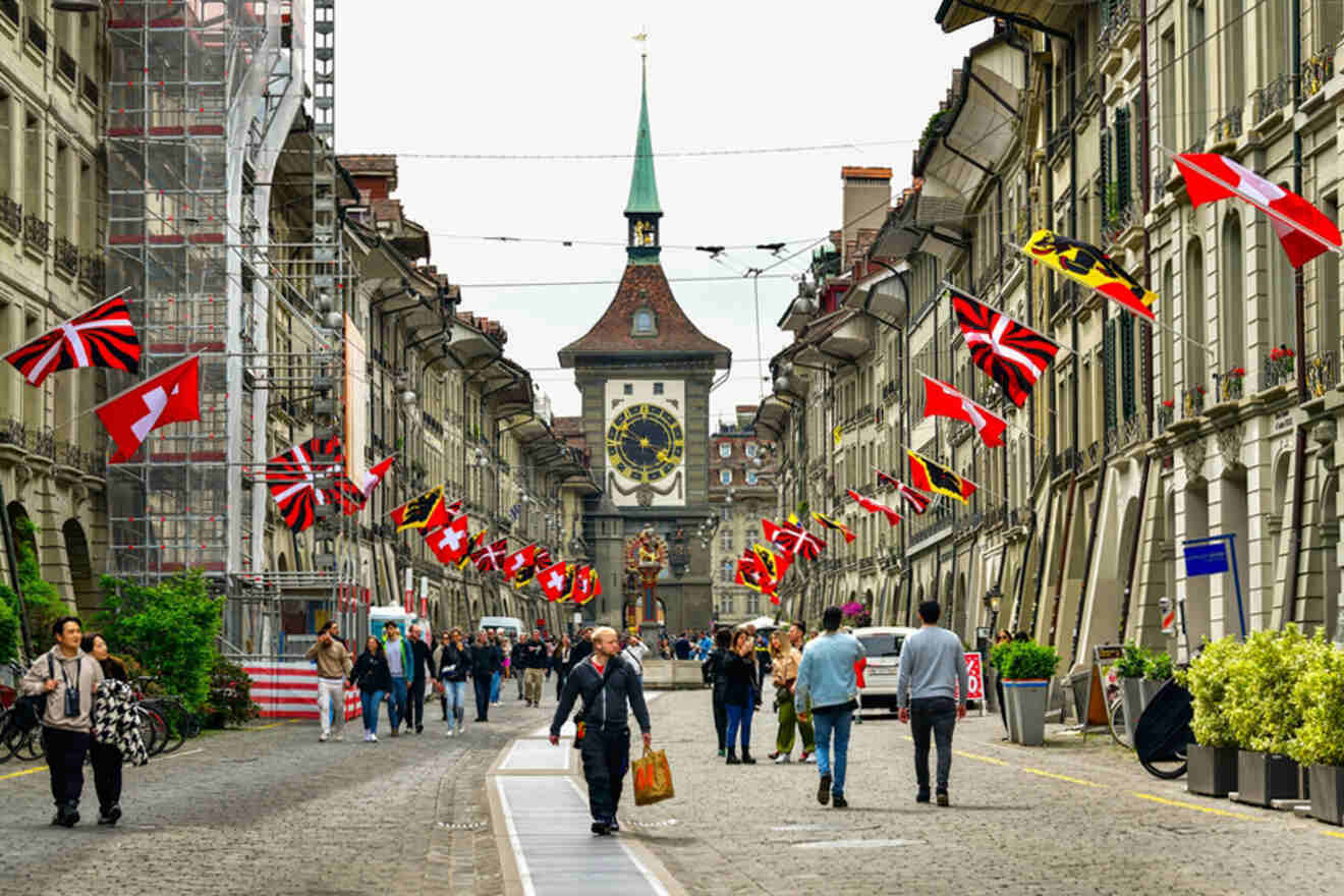 People walk along a cobblestone street lined with historic buildings and flags. A clock tower is seen in the background. Scaffolding and potted plants are present on the sides of the street.