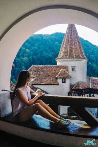 Yulia, the founder of this website, sits on a ledge inside a circular window, looking out at a medieval tower with a steep, conical roof, surrounded by forested hills.