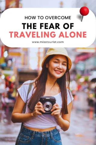 A woman holds a camera and smiles in a busy street. Text above reads, "How to Overcome the Fear of Traveling Alone." Website: www.misstourist.com.