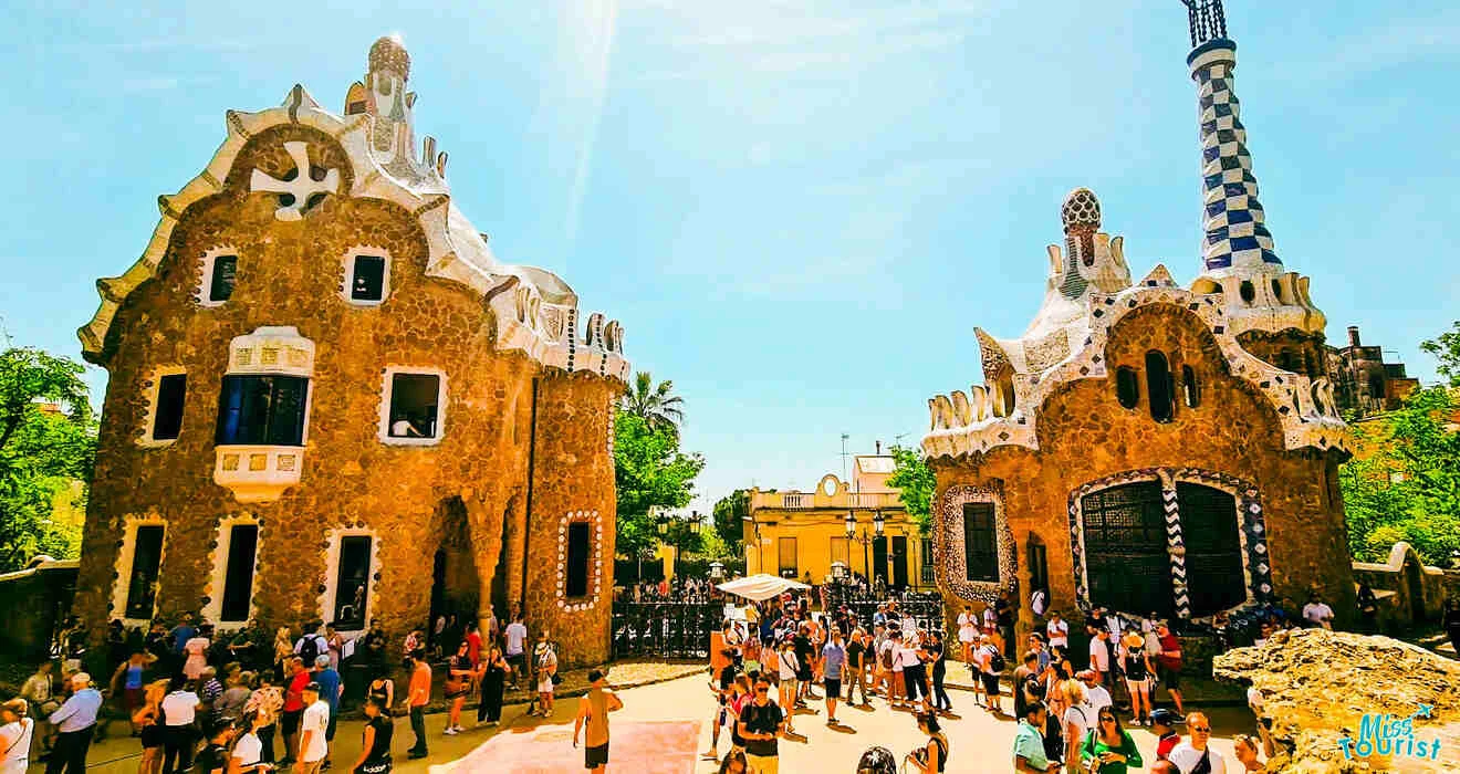 Crowd of people visiting the iconic entrance of Park Güell in Barcelona, featuring two uniquely designed buildings with intricate architectural details.