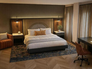 Modern hotel room with a large bed, two nightstands, pendant lights, an armchair, a desk with a chair, a rug, and floor-to-ceiling curtains.