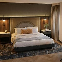 A modern hotel room with a king bed, two nightstands with lamps, a decorative headboard, and a patterned rug.