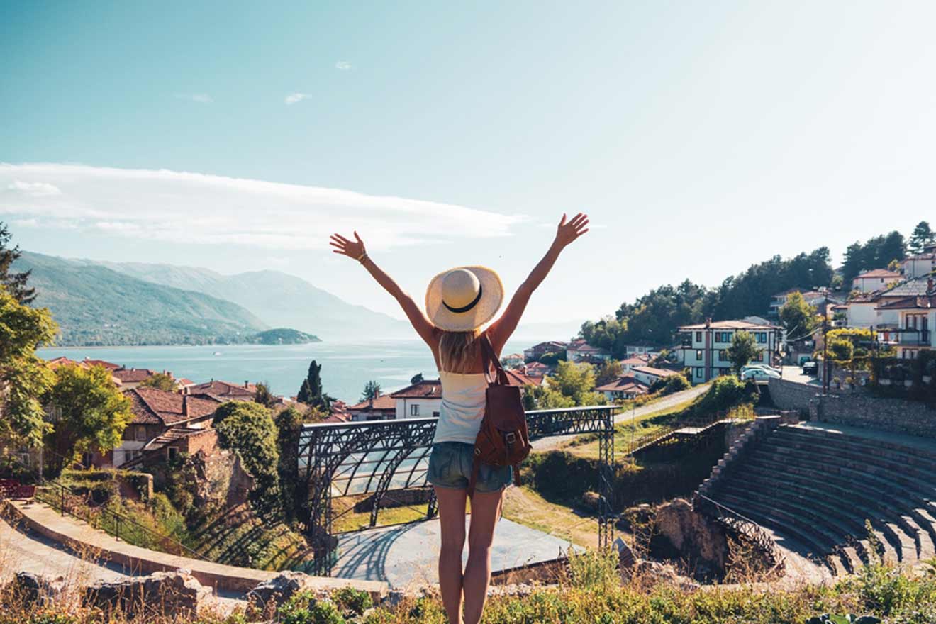 A person wearing a hat stands with arms raised, overlooking a scenic lake, town, and amphitheater.