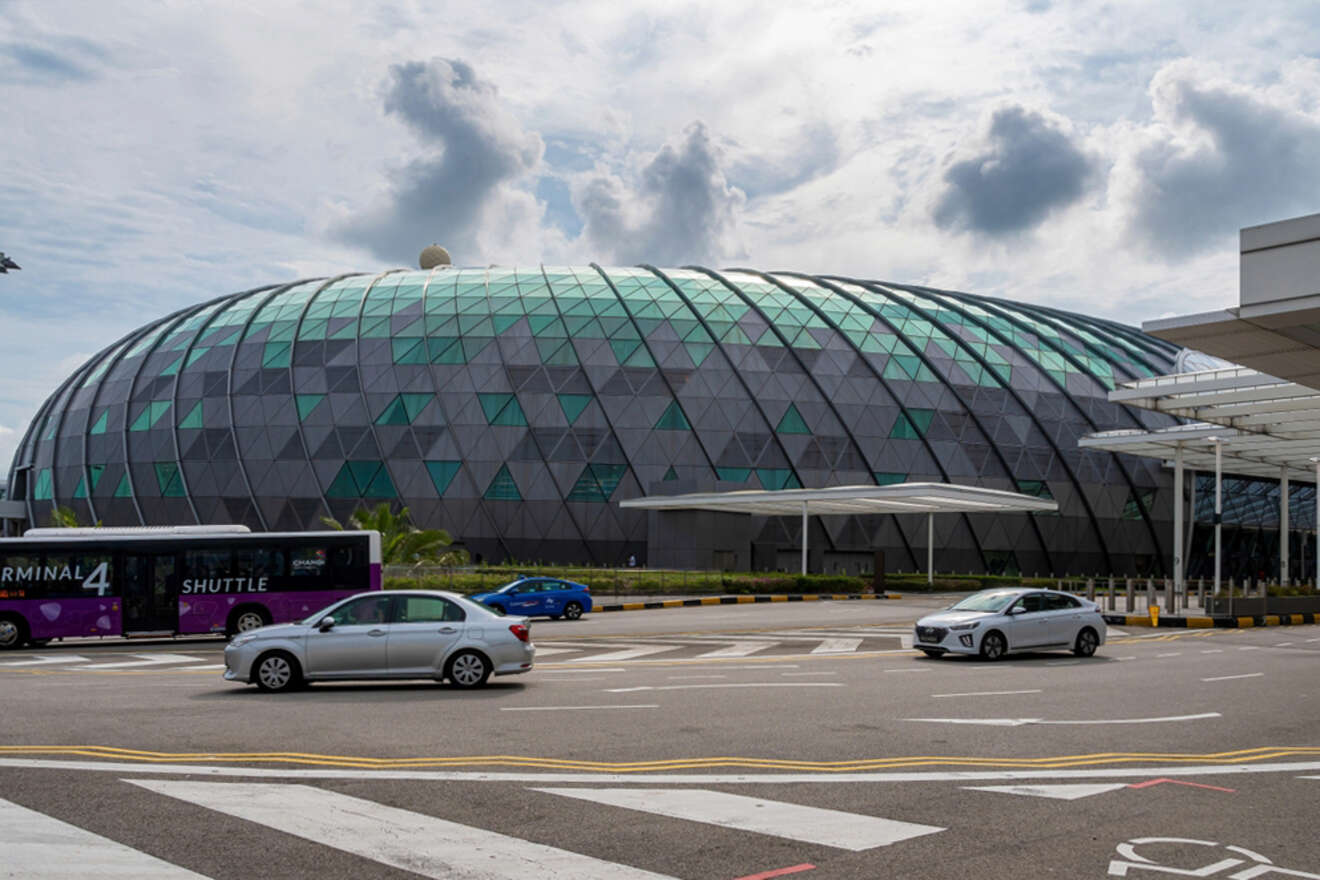 Large domed building with geometric glass and metal pattern, situated behind a road with cars passing by. Shuttle bus visible on the left. Cloudy sky overhead.