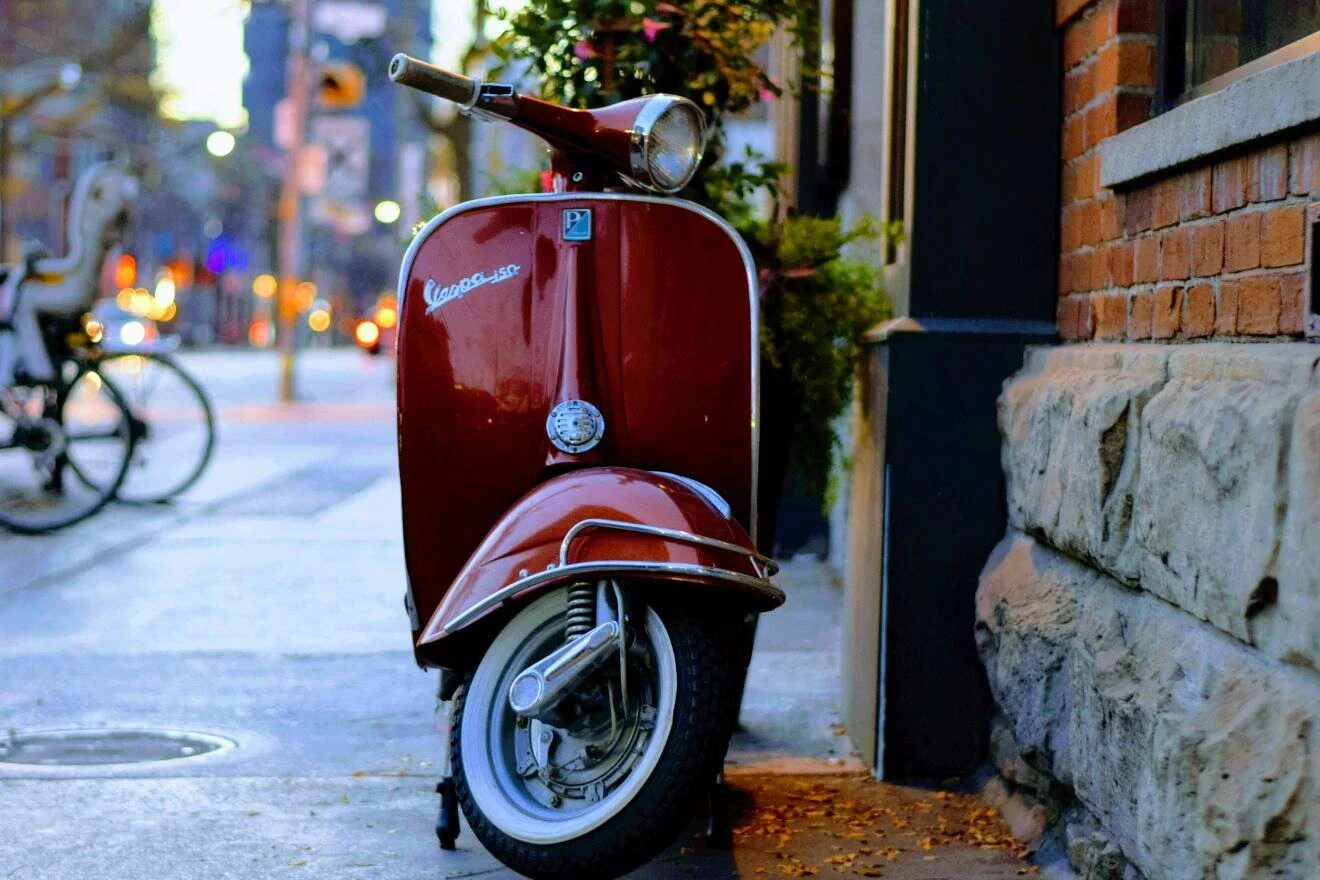 A red Vespa scooter parked on a city sidewalk near a brick wall, with a blurred background of street and bicycle.