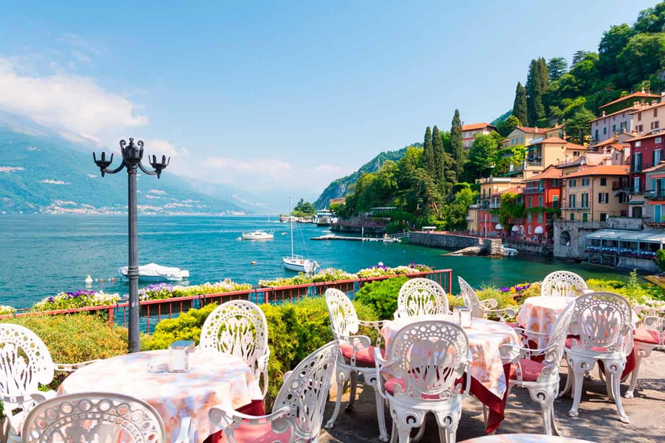 Outdoor café with colorful tables and chairs, overlooking a serene lake with boats, surrounded by lush hills and colorful buildings.