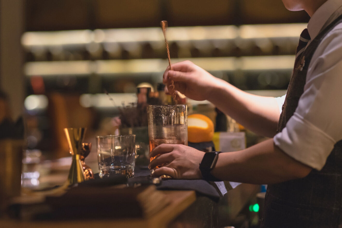 A bartender stirs a cocktail in a dimly lit bar, surrounded by various tools and glassware on the counter