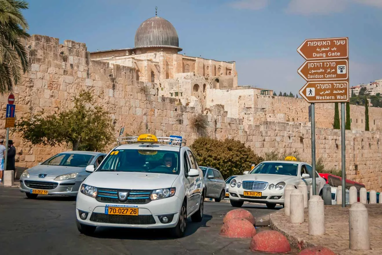 Several taxis wait near the entrance of the Dung Gate in Jerusalem, with directional signs for the Dung Gate, Zion Gate, and Western Wall visible. Stone walls and a domed building are in the background.